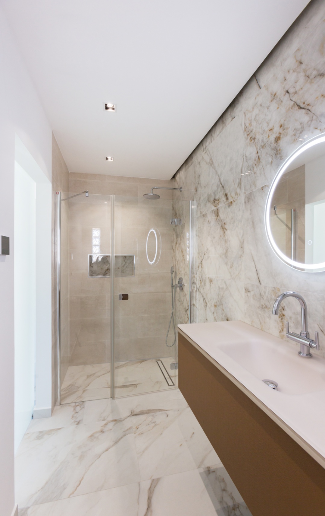 Salle de bain moderne avec douche à l'italienne vitrée, carrelage effet marbre, meuble vasque suspendu et miroir circulaire à rétroéclairage LED intégré.