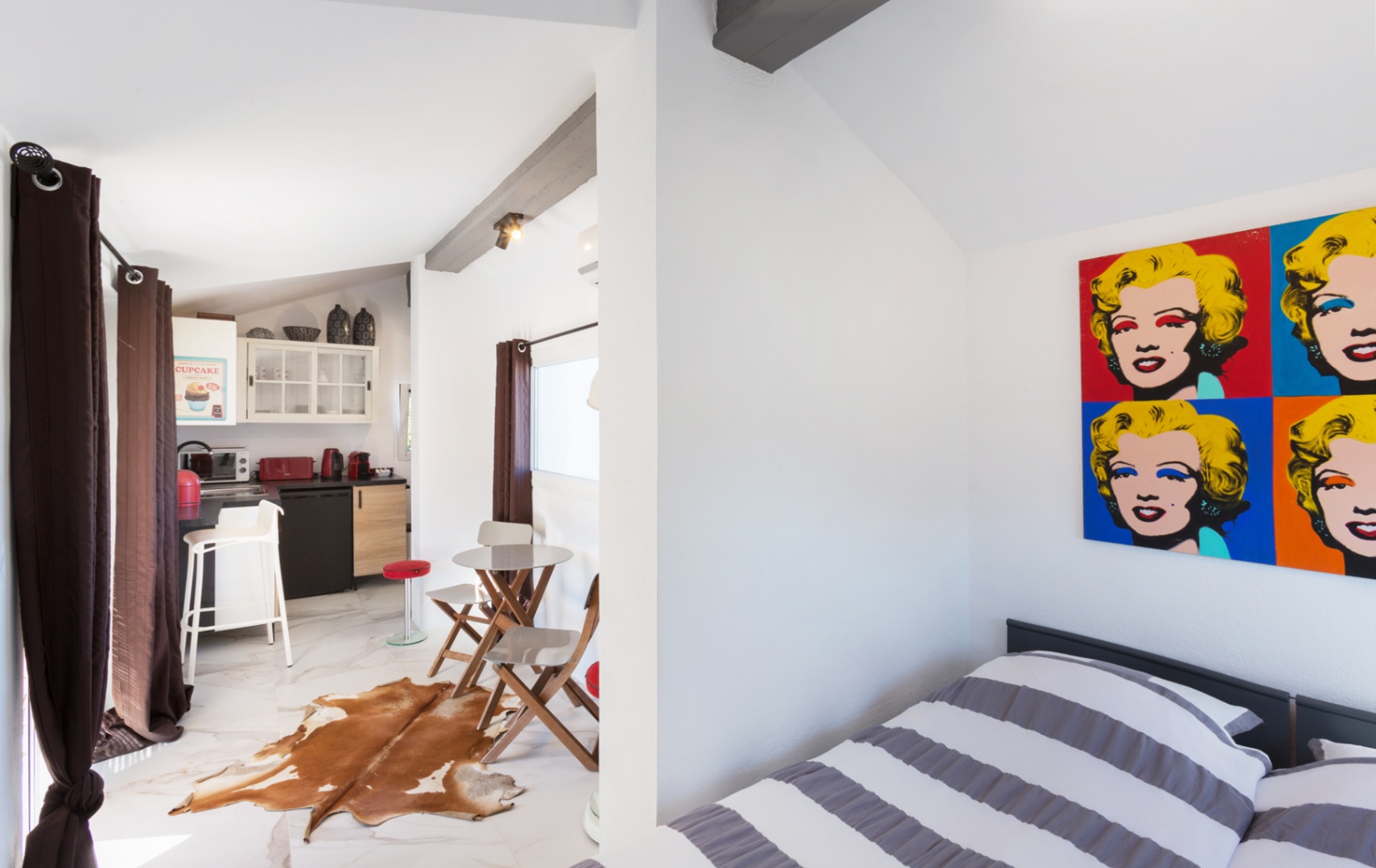 Studio avec décor pop-art, poutres apparentes et sol effet marbre. Comprend une kitchenette avec appareils rouges et un coin repas avec tapis en peau de vache.