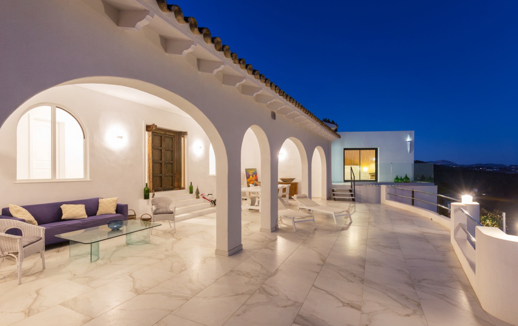 Terrasse couverte au crépuscule avec arcades blanches, sol en grès cérame effet marbre, salon extérieur et chaises longues face à la vallée.