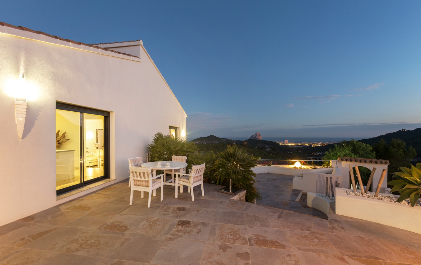 Terrasse en pierre naturelle avec mobilier de jardin blanc, offrant une vue panoramique sur le littoral de Calpe et le Peñón de Ifach au crépuscule.