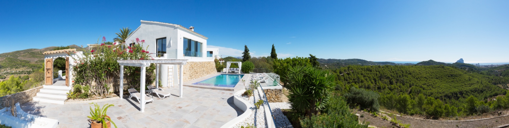 Terrasse panoramique avec piscine, pergola blanche et dallage en pierre, offrant une vue dégagée sur la vallée verdoyante et la mer Méditerranée au loin.