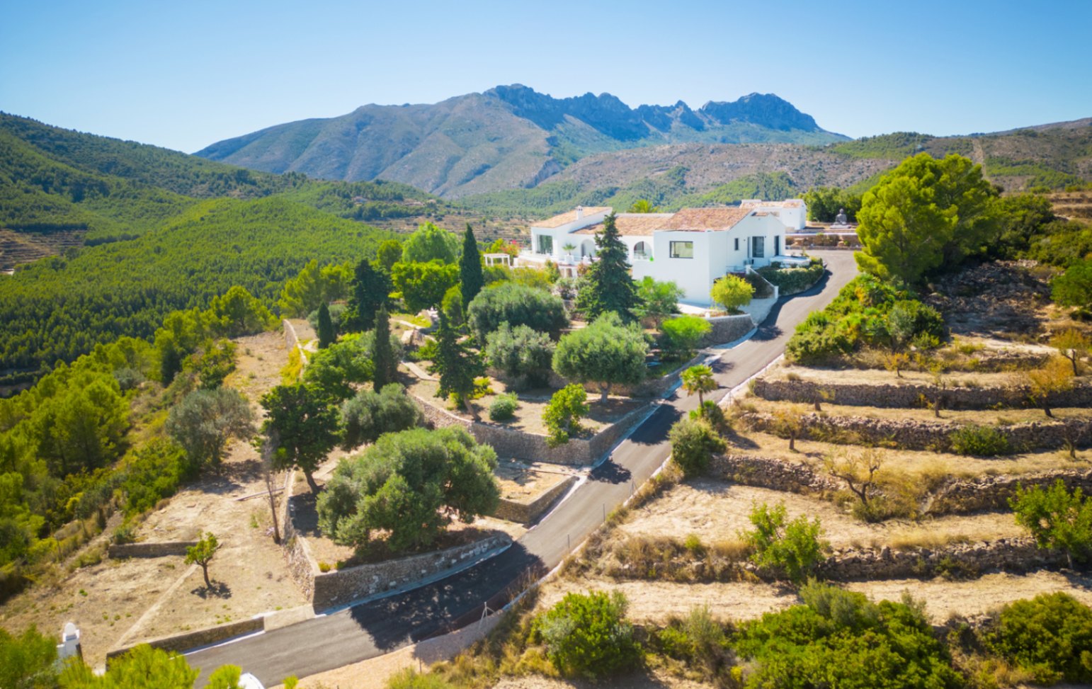 Vue aérienne d'une villa blanche nichée dans un paysage montagneux avec terrasses en pierre, jardins méditerranéens et vue panoramique sur la forêt de pins.