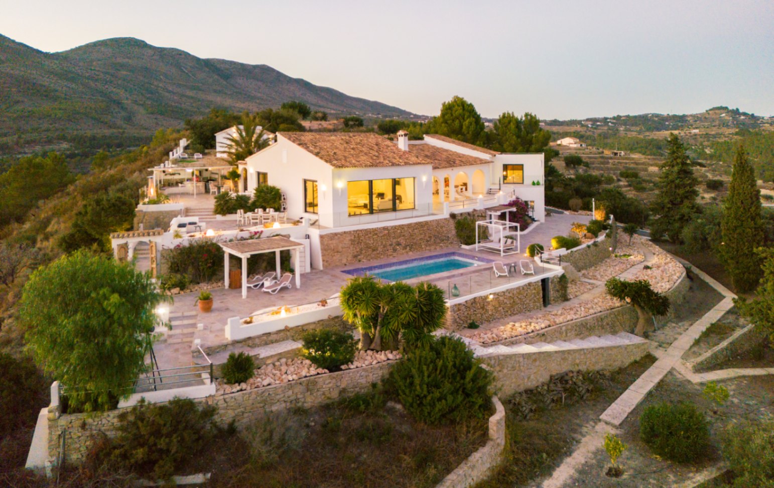 Vue aérienne d'une villa méditerranéenne aux façades blanches, toits en terre cuite et terrasses à plusieurs niveaux avec piscine et murs en pierre sèche.