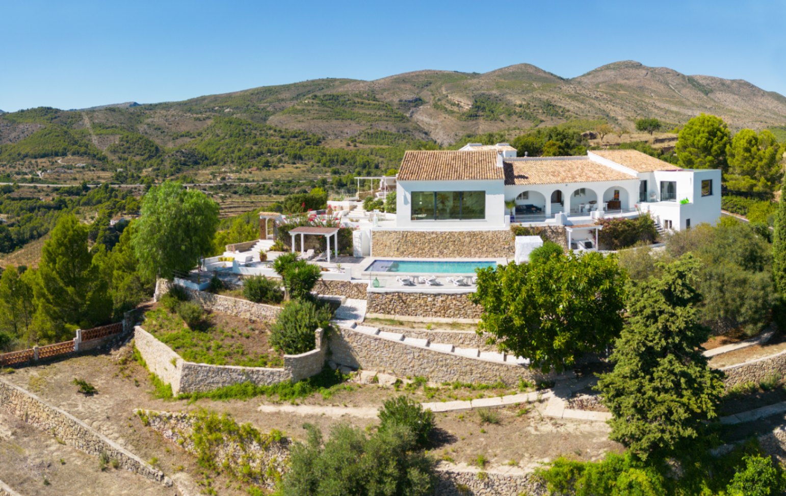 Vue aérienne d'une villa méditerranéenne blanche avec jardins en terrasses de pierre, piscine privée et porche à arcades dominant le paysage montagneux.