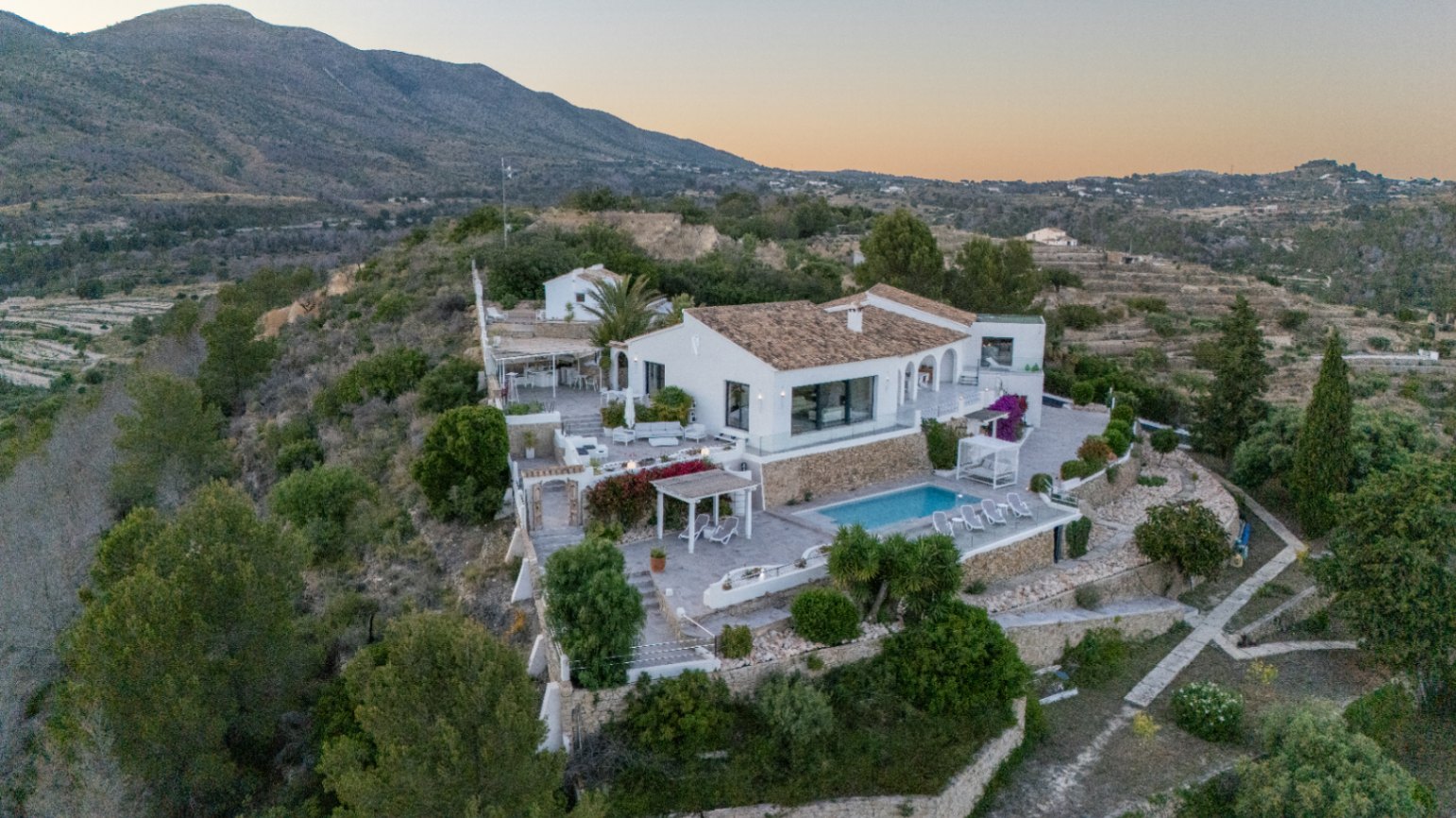 Vue surélevée d'une villa méditerranéenne blanche avec murs en pierre, piscine et plusieurs terrasses aménagées sur fond de montagnes.