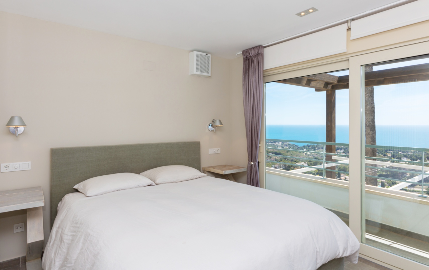 Chambre avec tête de lit en tissu vert et grandes baies vitrées ouvrant sur une terrasse avec pergola en bois et vue panoramique sur la mer.