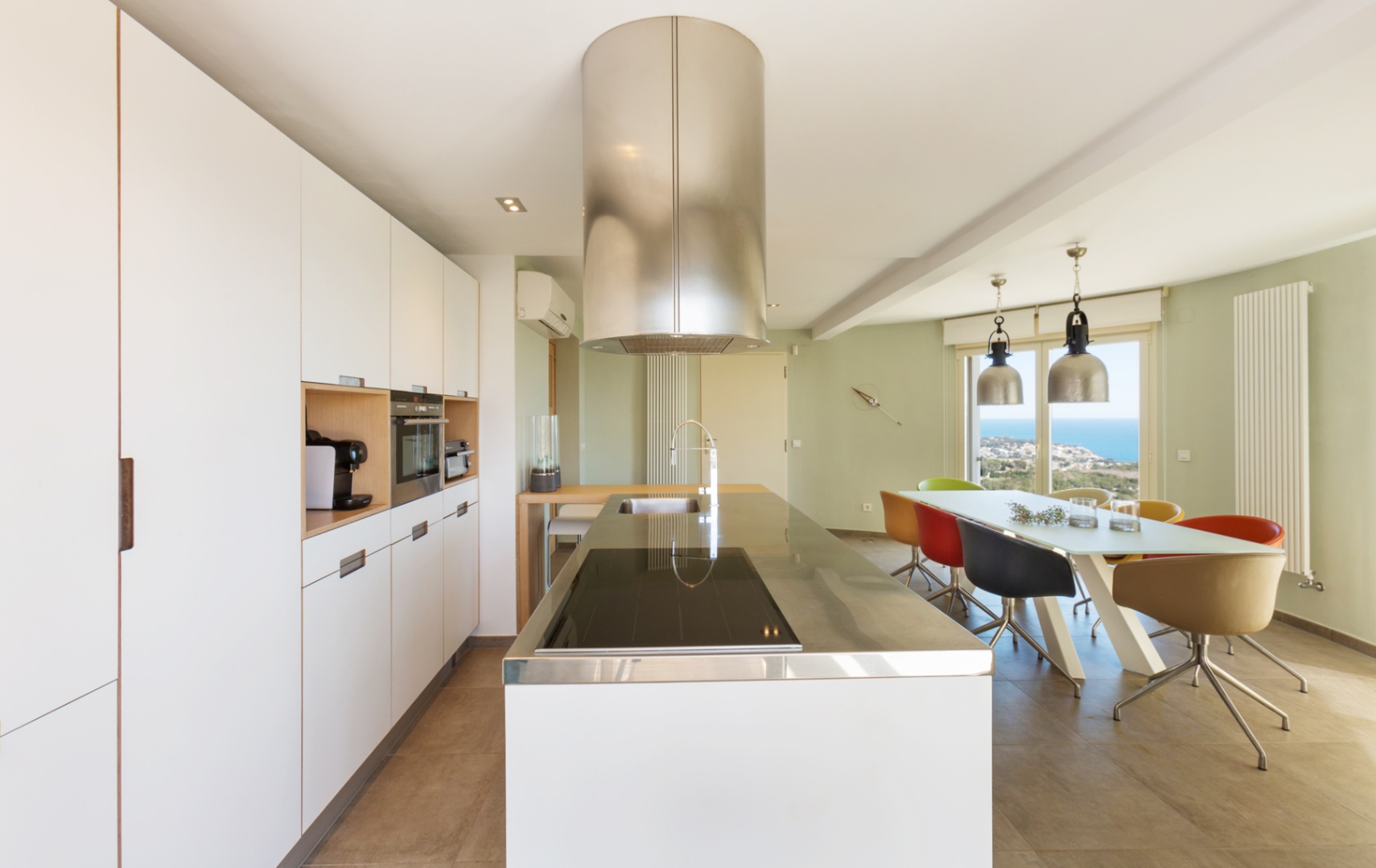 Cuisine moderne ouverte avec îlot blanc, plan de travail en inox, hotte cylindrique et coin repas offrant une vue sur la mer par de grandes baies vitrées.