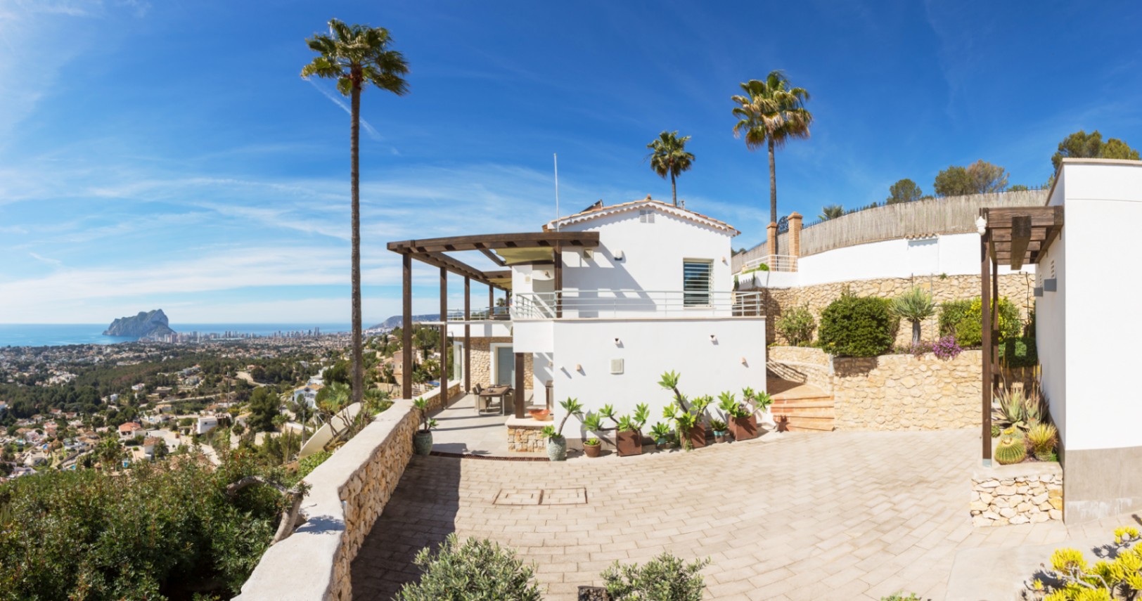 Extérieur d'une villa méditerranéenne avec façades blanches, pergolas en bois et murs en pierre, offrant une vue panoramique sur la côte et le Peñón de Ifach.