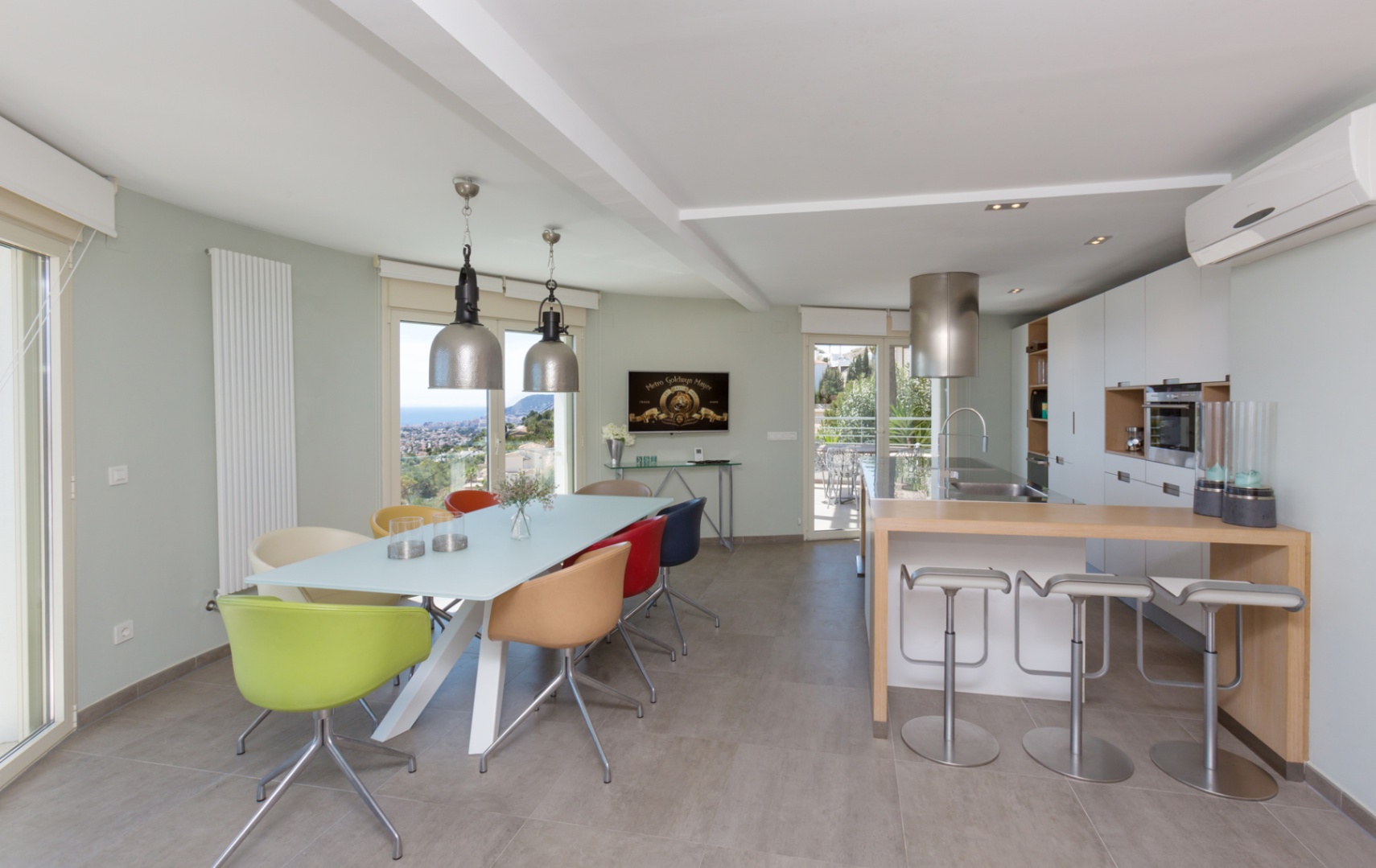 Intérieur à aire ouverte avec cuisine moderne, comptoir en bois et salle à manger aux chaises colorées sous des suspensions industrielles, offrant une vue côtière.