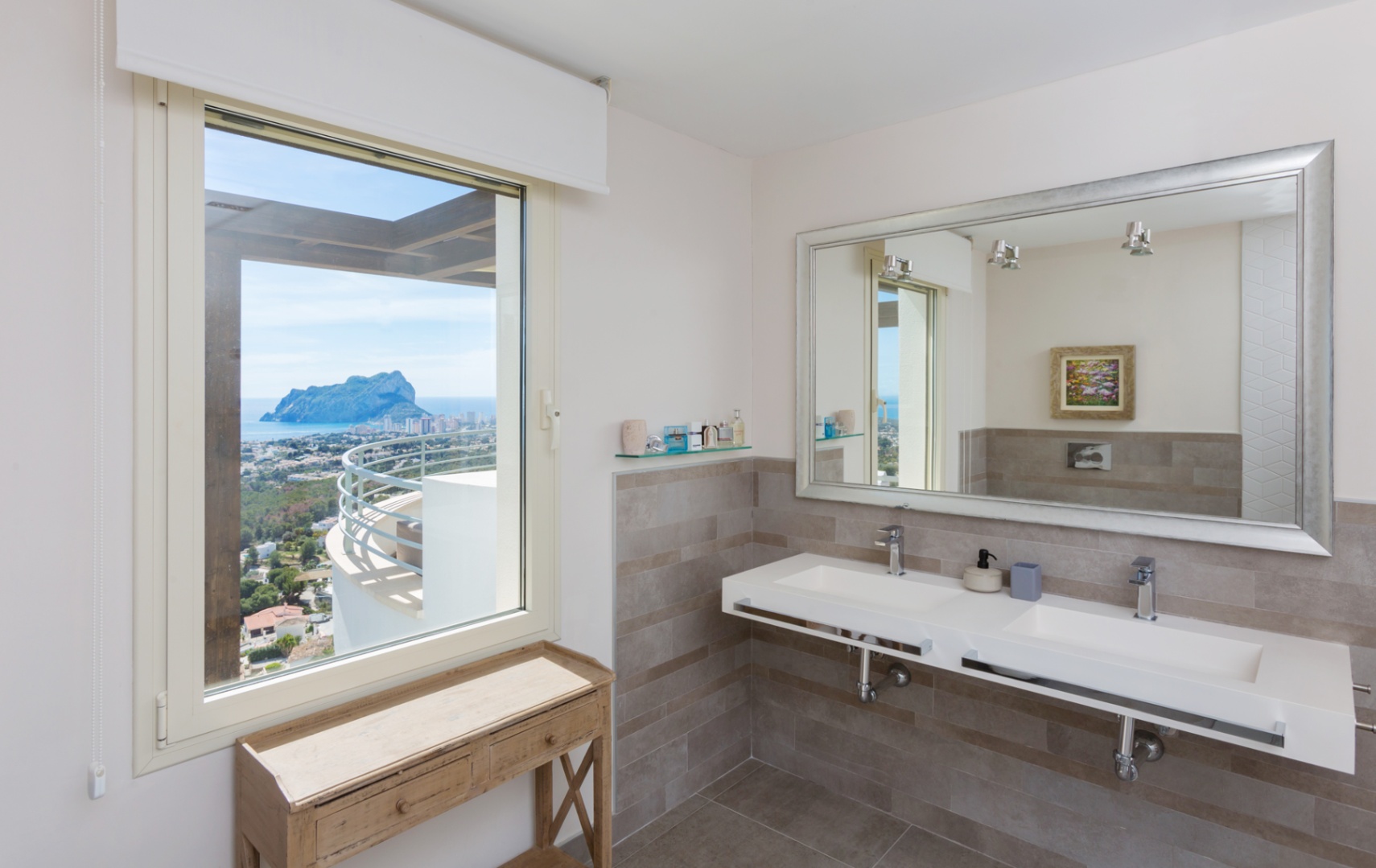 Salle de bain moderne avec double vasque suspendue, grand miroir au cadre argenté et fenêtre offrant une vue panoramique sur le Peñón de Ifach et la mer.
