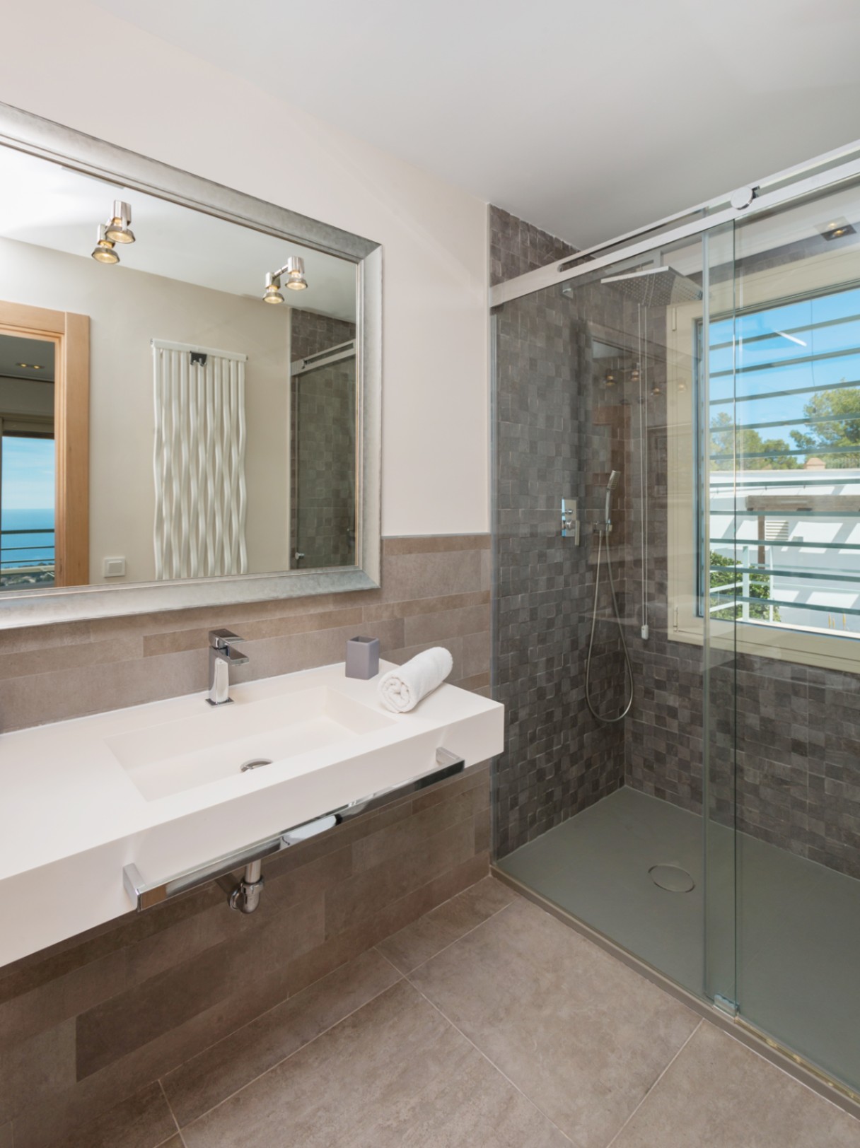 Salle de bain moderne avec vasque intégrée blanche, grand miroir à cadre argenté et douche à l'italienne avec mosaïque grise et lumière naturelle.