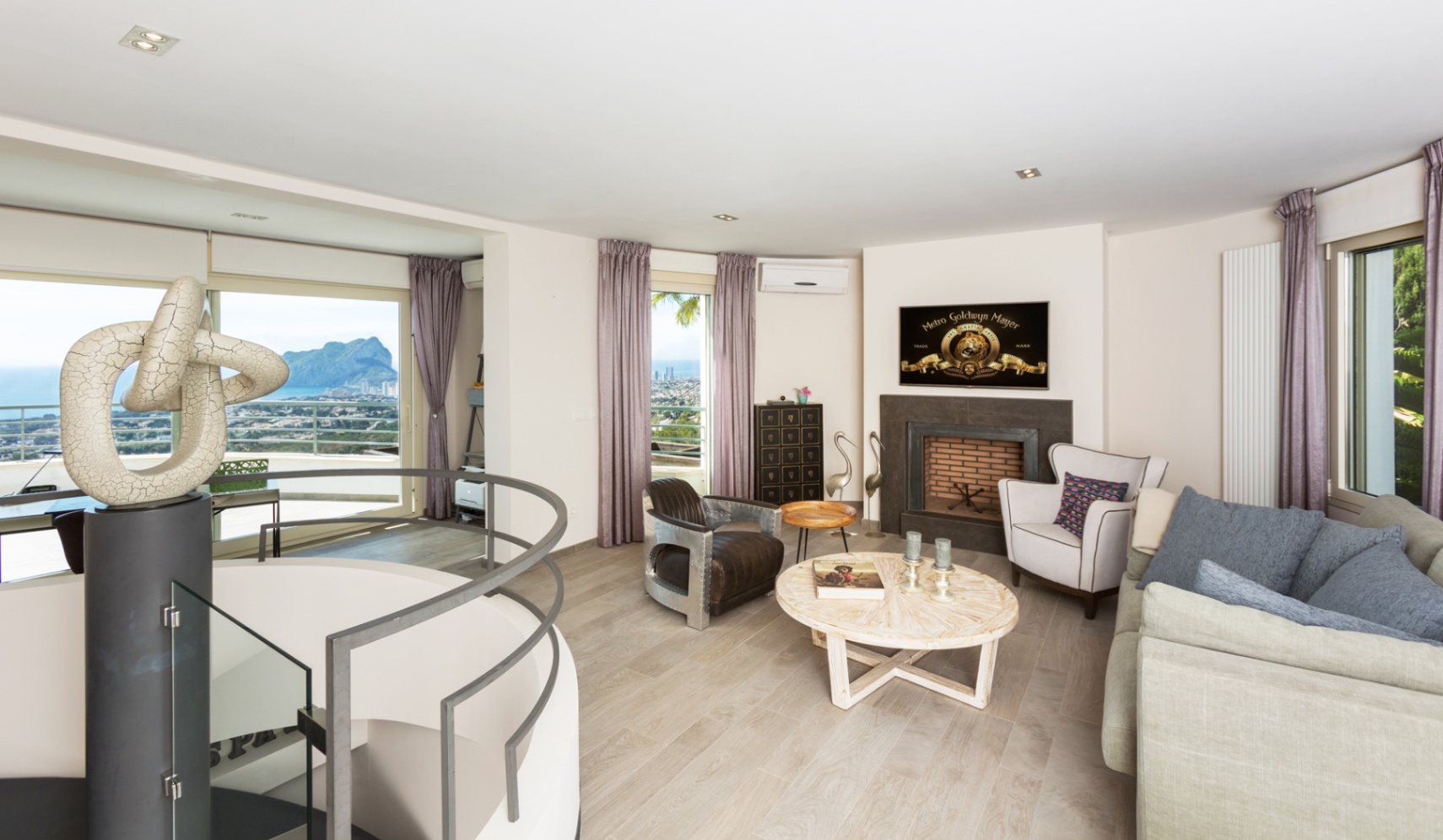 Salon moderne comprenant une cheminée, un escalier en colimaçon avec sculpture et de grandes baies vitrées donnant sur la mer et le Peñón de Ifach.