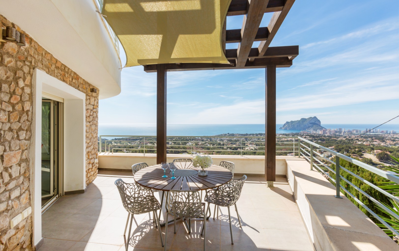 Terrasse avec murs en pierre naturelle, pergola en bois et voile d'ombrage. Espace repas extérieur offrant une vue panoramique sur la Méditerranée et le Peñón de Ifach.