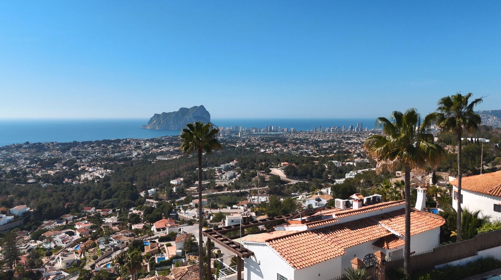Vue panoramique sur le Peñón de Ifach et la ville de Calpe, avec des villas méditerranéennes aux toits de tuiles et des palmiers au premier plan.