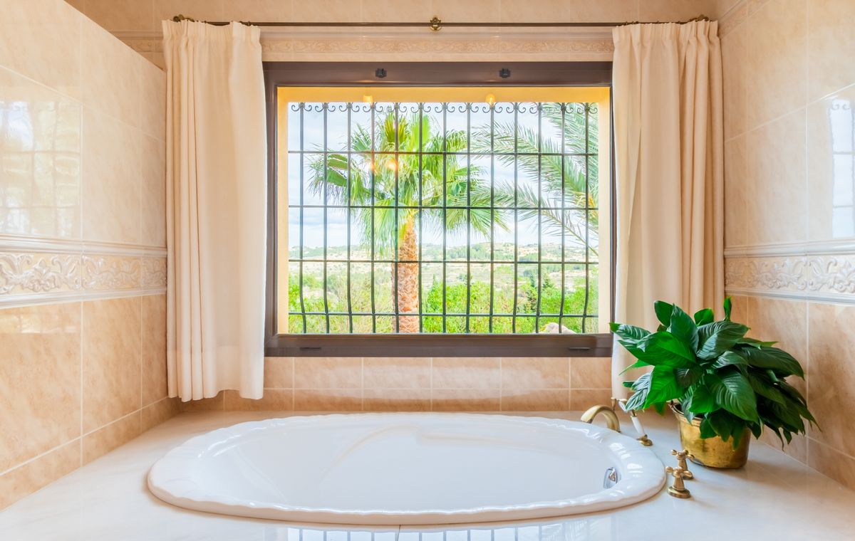 Baignoire ovale encastrée avec robinetterie dorée, située sous une fenêtre avec ferronnerie offrant une vue sur les palmiers et la campagne.