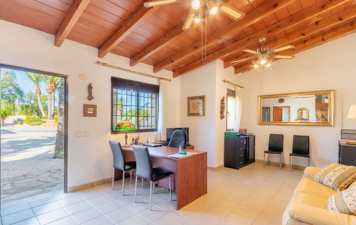 Bureau avec plafond à poutres apparentes, ventilateurs de plafond et mobilier en bois. Une porte ouverte laisse entrer la lumière naturelle et offre une vue sur le jardin.