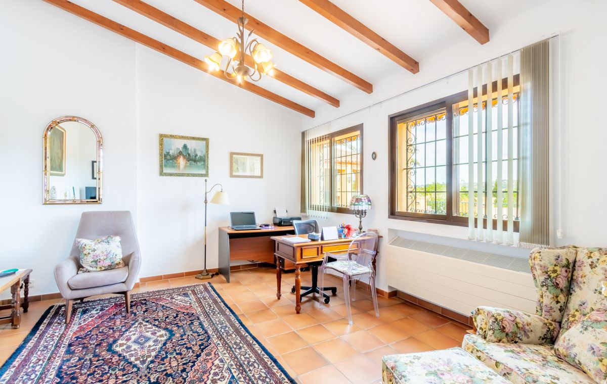 Bureau avec poutres en bois apparentes, sol en terre cuite et deux espaces de travail. Grandes fenêtres avec grilles de sécurité offrant une lumière naturelle.