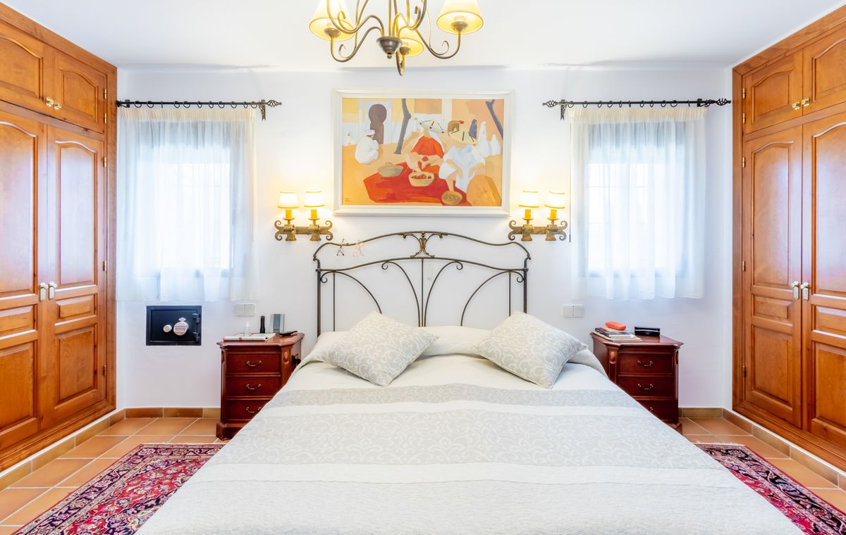 Chambre avec tête de lit en fer forgé, armoires encastrées en bois symétriques et sol en terre cuite. Comprend des appliques murales et un coffre-fort intégré.