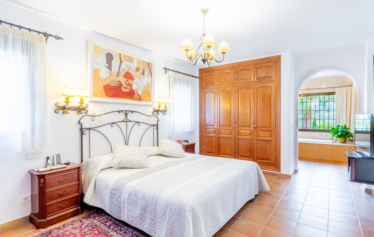 Chambre avec tête de lit en fer forgé, grands placards en bois et sol en terre cuite. Une arche s'ouvre sur une salle de bain attenante avec baignoire.