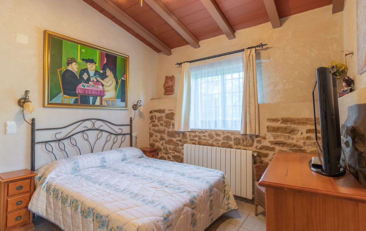 Chambre rustique avec lit en fer forgé, murs en pierres apparentes et plafond à poutres en bois. Un grand tableau figuratif et une commode en bois complètent la pièce.