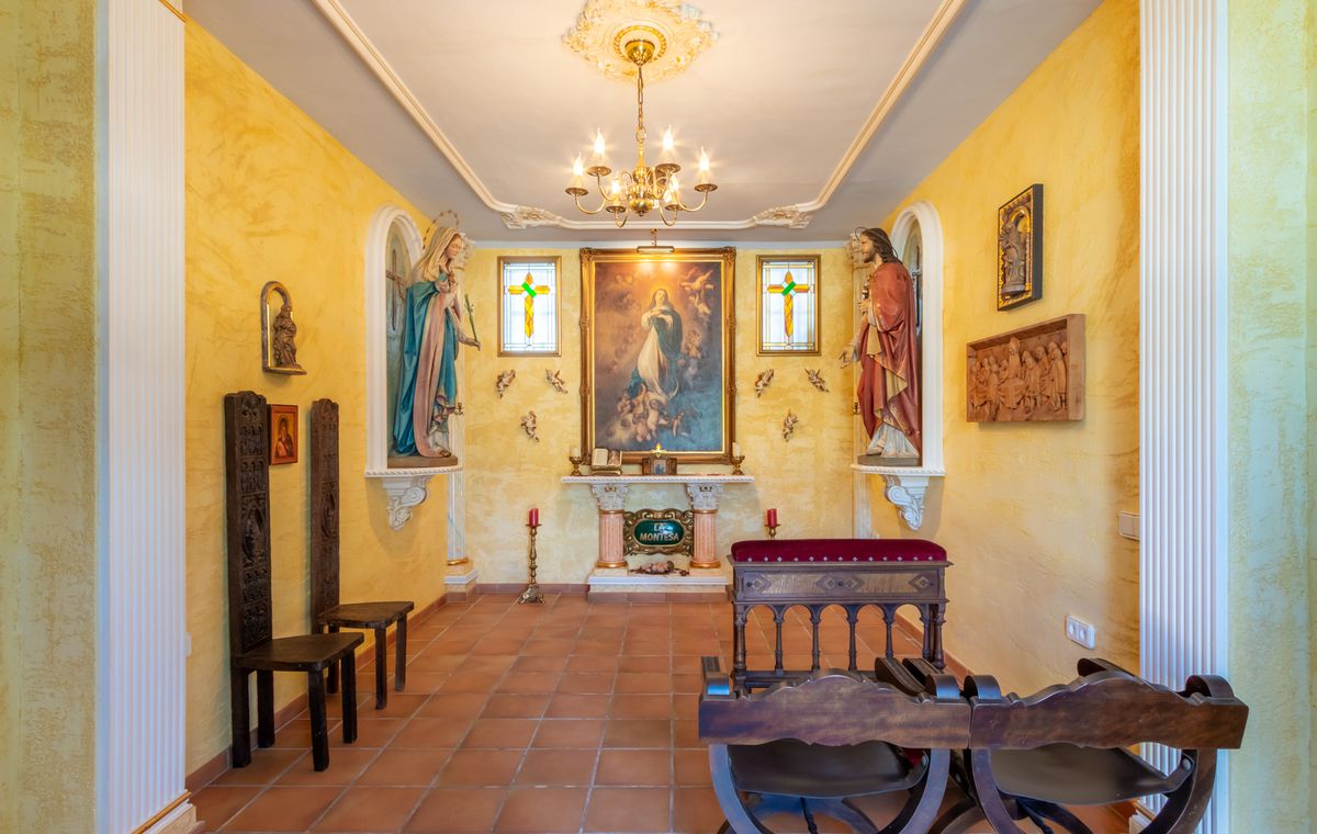 Chapelle privée avec murs texturés jaunes, sol en terre cuite, autel central orné de peintures religieuses, statues dans des niches et bancs en bois sculpté.