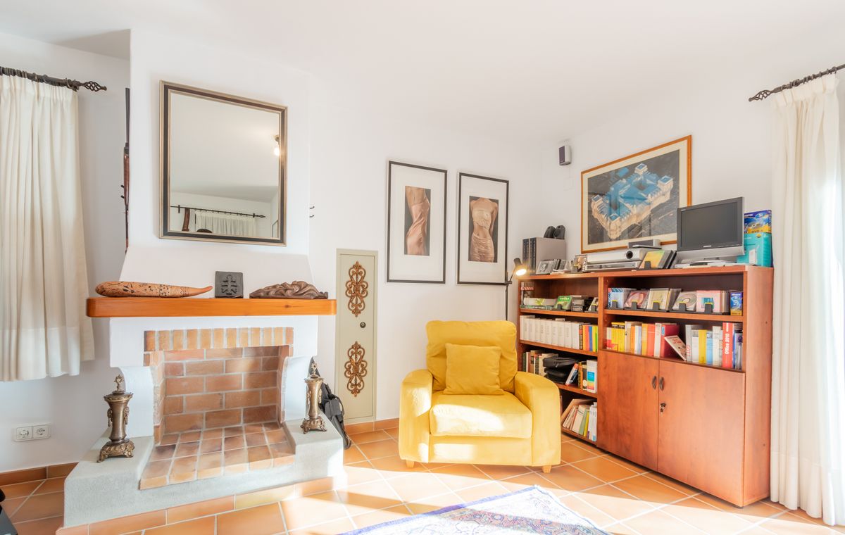 Coin salon avec cheminée en briques, fauteuil jaune et bibliothèque en bois sur un sol en terre cuite classique.
