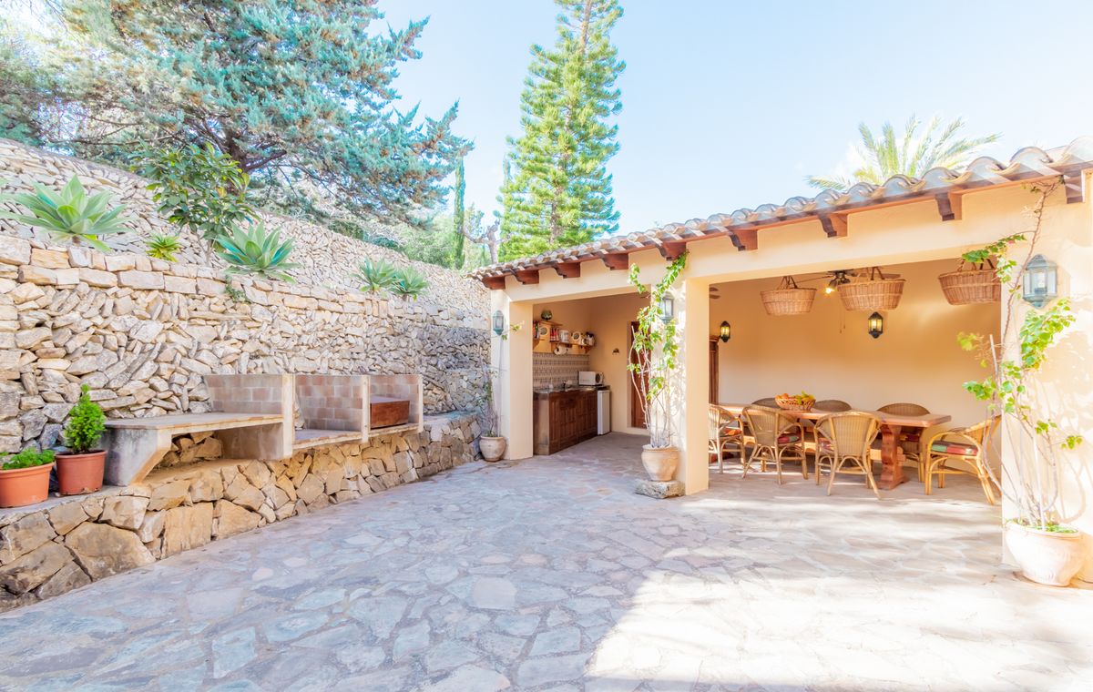 Espace repas extérieur avec terrasse en pierre, barbecue intégré et porche couvert aux poutres apparentes, comprenant une cuisine d'été et du mobilier en rotin.