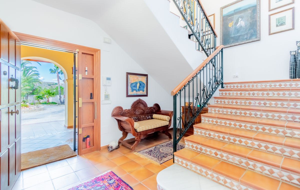 Hall d'entrée avec escalier en terre cuite aux contremarches décorées, rampe en fer forgé et banc en bois sculpté. La porte ouverte offre une vue sur le patio ensoleillé.