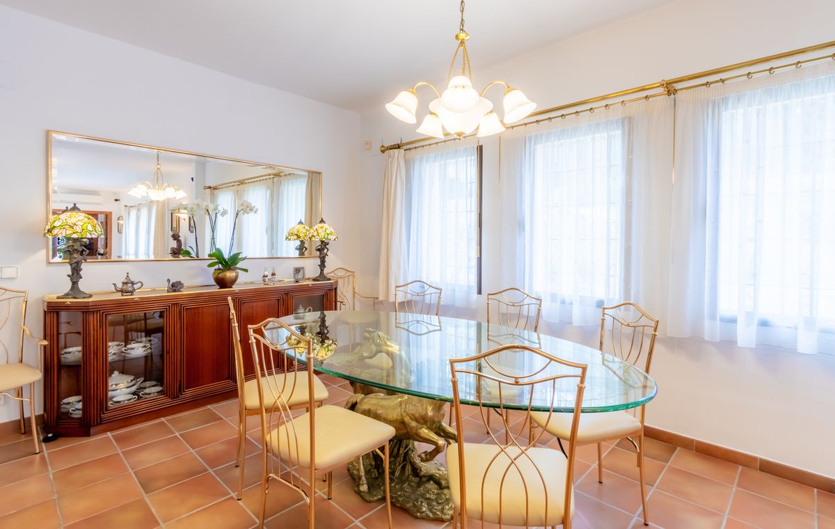 Salle à manger formelle avec table ovale en verre sur socle sculpté en laiton, chaises dorées, sol en terre cuite et buffet en bois avec miroir.