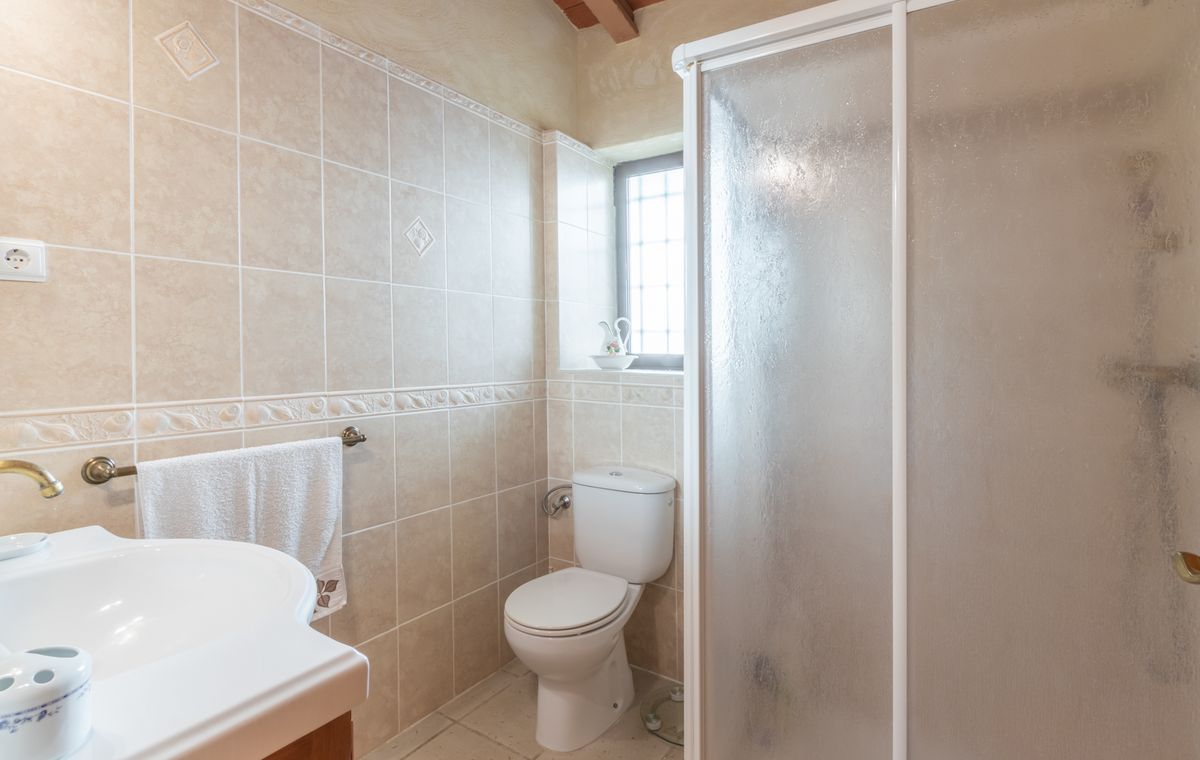 Salle de bain avec carrelage beige à motifs, toilettes blanches et cabine de douche givrée, complétée par des poutres en bois apparentes au plafond.