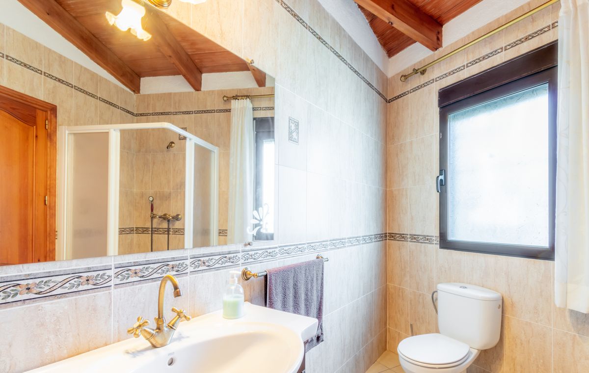 Salle de bain avec plafond en bois mansardé, poutres apparentes, carrelage beige à motifs et robinetterie en laiton de style ancien.