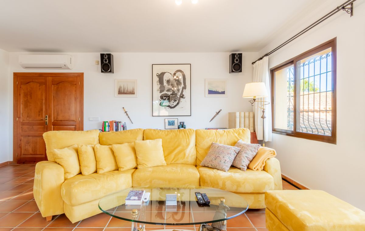 Salon avec un grand canapé d'angle jaune, une table basse en verre et un sol en terre cuite. Les murs blancs sont ornés d'œuvres d'art et de haut-parleurs muraux.