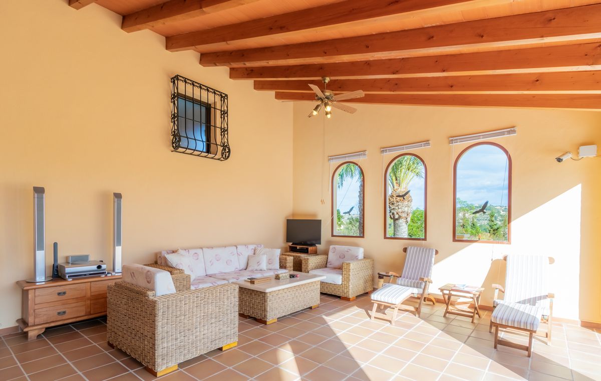 Salon de jardin avec poutres apparentes, sol en terre cuite et mobilier en osier. Trois fenêtres cintrées offrent une vue sur les palmiers environnants.
