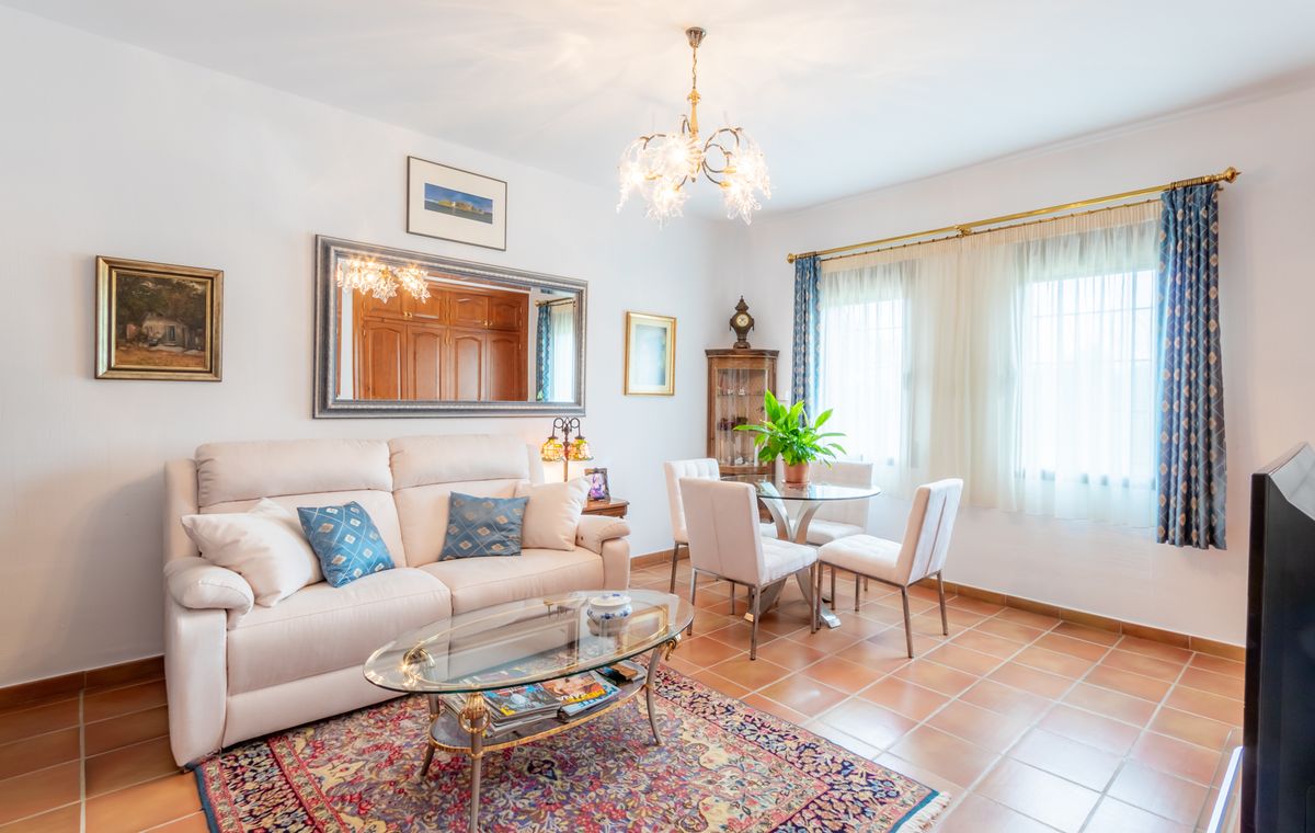 Salon lumineux avec sol en terre cuite, canapé blanc agrémenté de coussins bleus et coin repas avec table en verre sous un lustre décoratif.