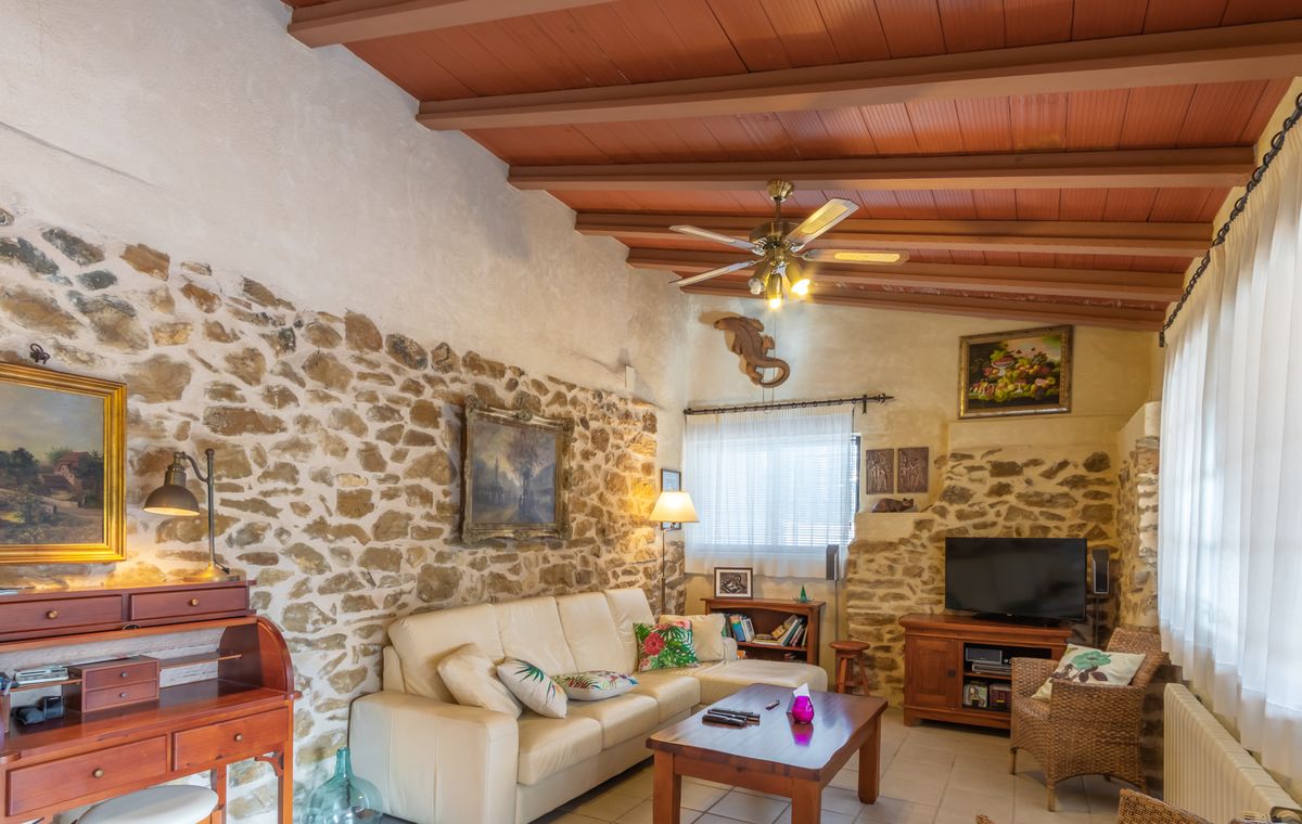 Salon rustique avec murs en pierre apparente, plafond à poutres en bois et canapé en cuir crème, agrémenté d'un bureau secrétaire classique.