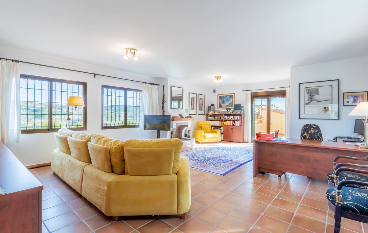 Salon spacieux avec sol en terre cuite, grand canapé d'angle jaune, espace bureau avec bureau en bois et cheminée.