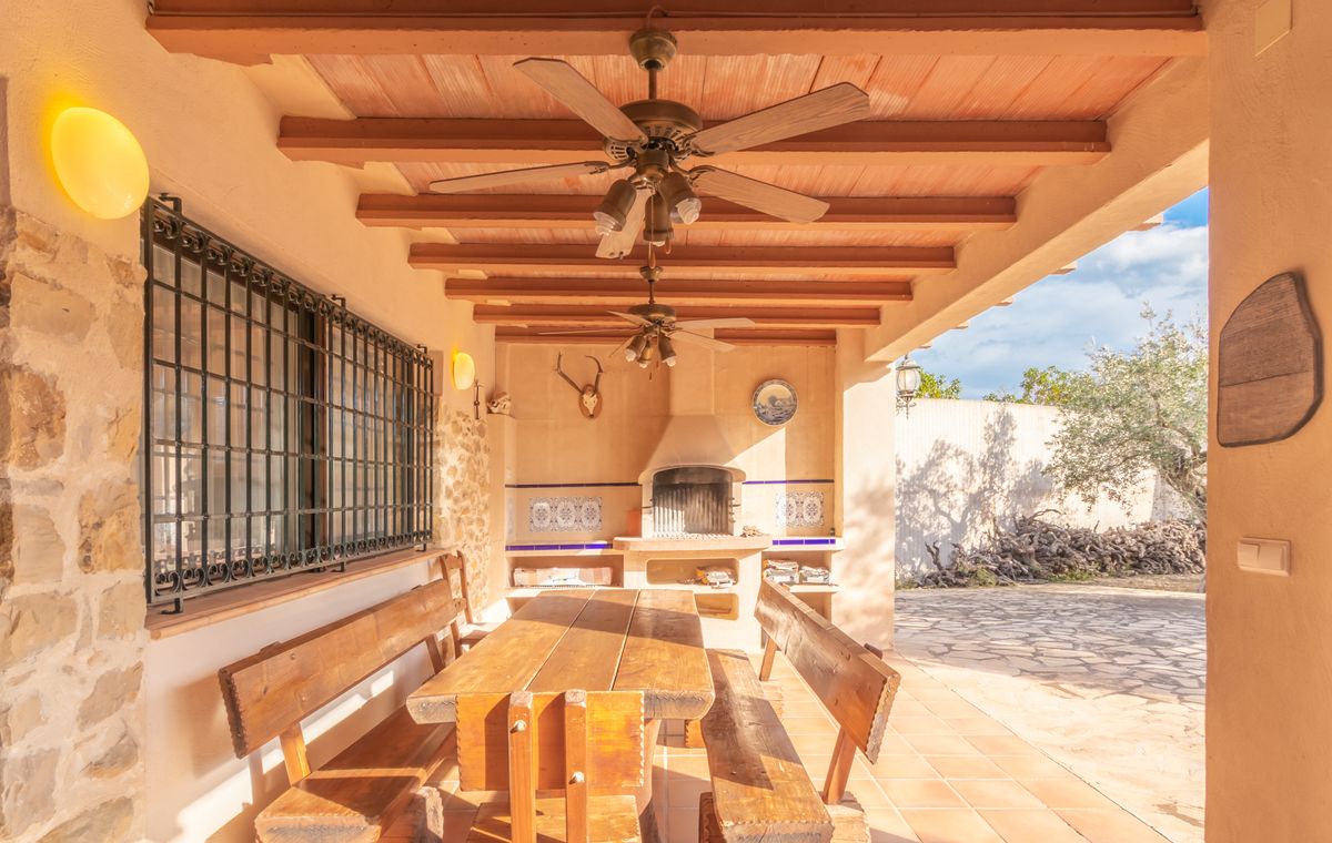 Terrasse couverte avec plafond à poutres en bois et ventilateurs, comprenant un coin repas rustique et un barbecue intégré orné de carreaux décoratifs.