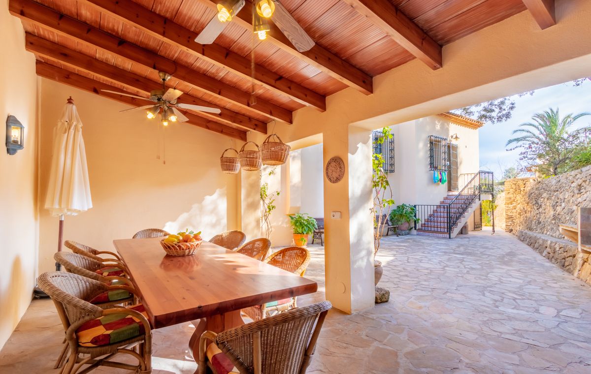 Terrasse couverte avec plafond en poutres de bois, ventilateurs et espace dînatoire en osier s'ouvrant sur une cour pavée de pierres.
