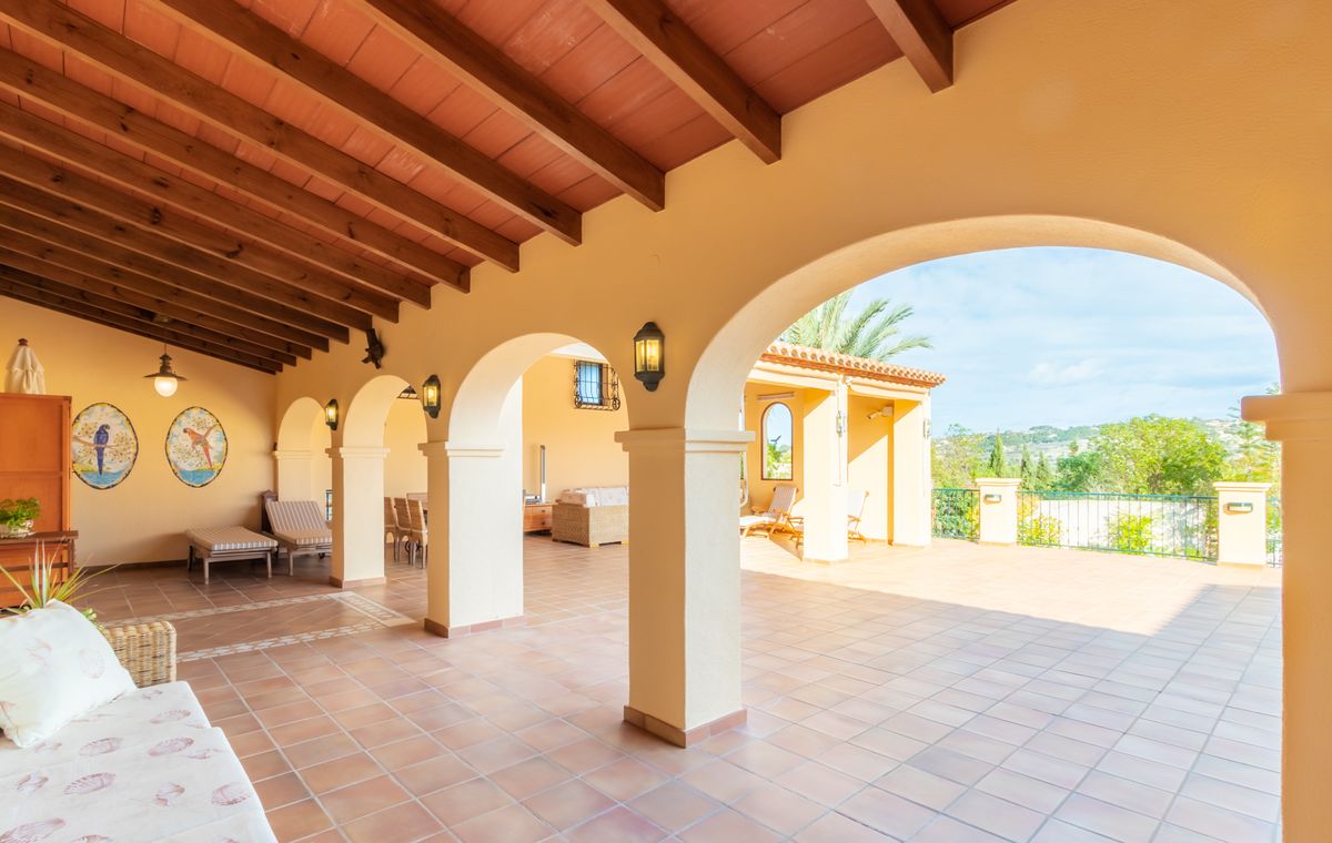 Terrasse couverte avec poutres en bois sombre, sol en terre cuite orné de frises et arcades s'ouvrant sur un vaste patio extérieur ensoleillé.