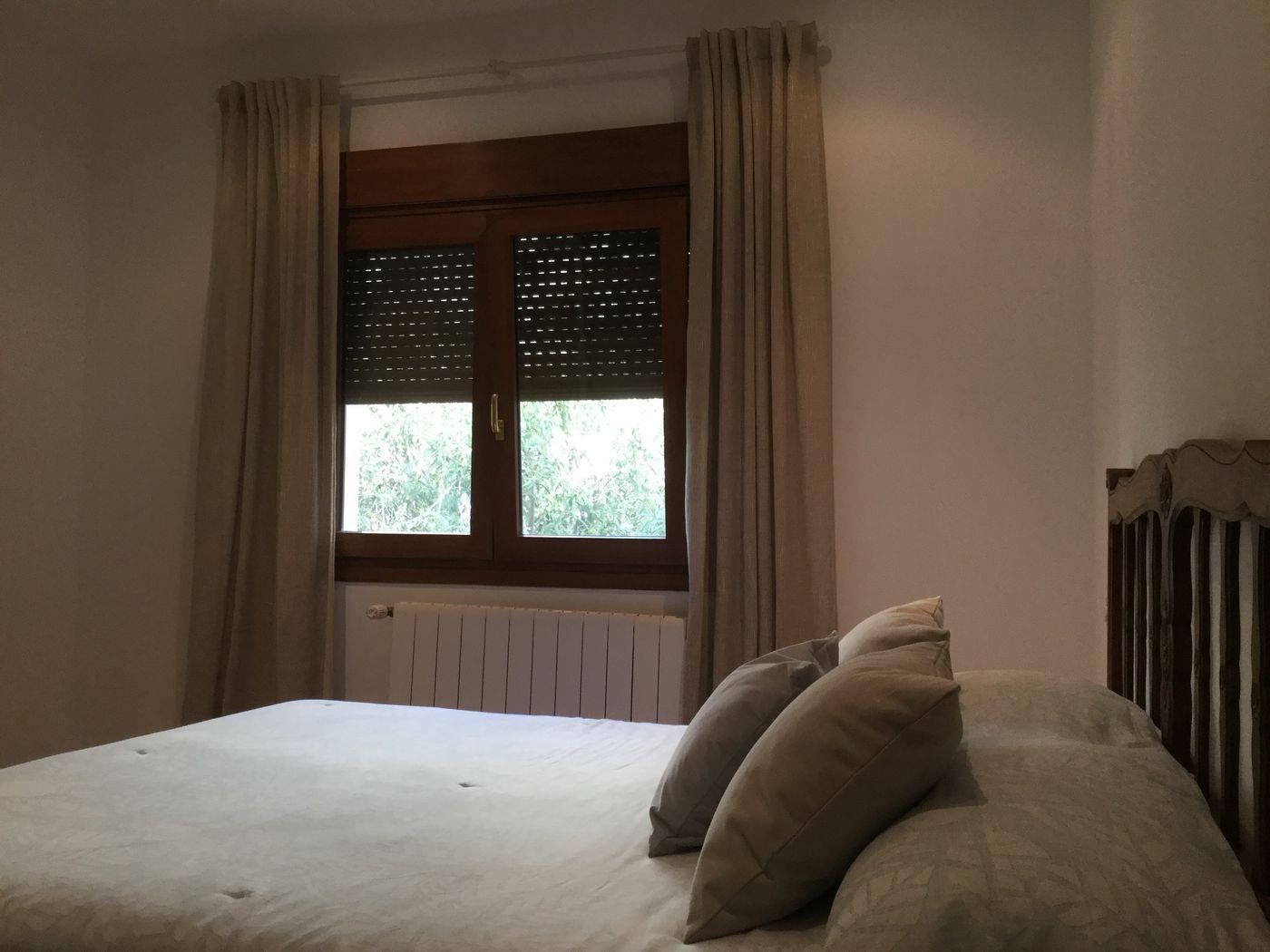 Chambre à coucher avec tête de lit en bois sculpté, fenêtre équipée de volets sombres, rideaux beiges et radiateur blanc mural.