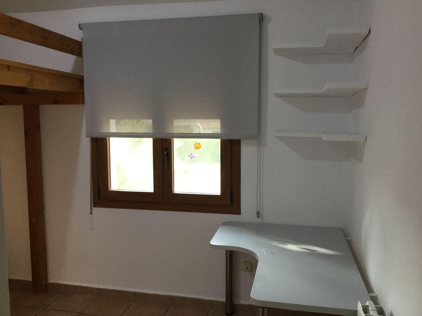Chambre avec lit mezzanine en bois, bureau d'angle et étagères murales blanches. Une fenêtre à cadre en bois avec store enroulable gris apporte de la clarté naturelle.