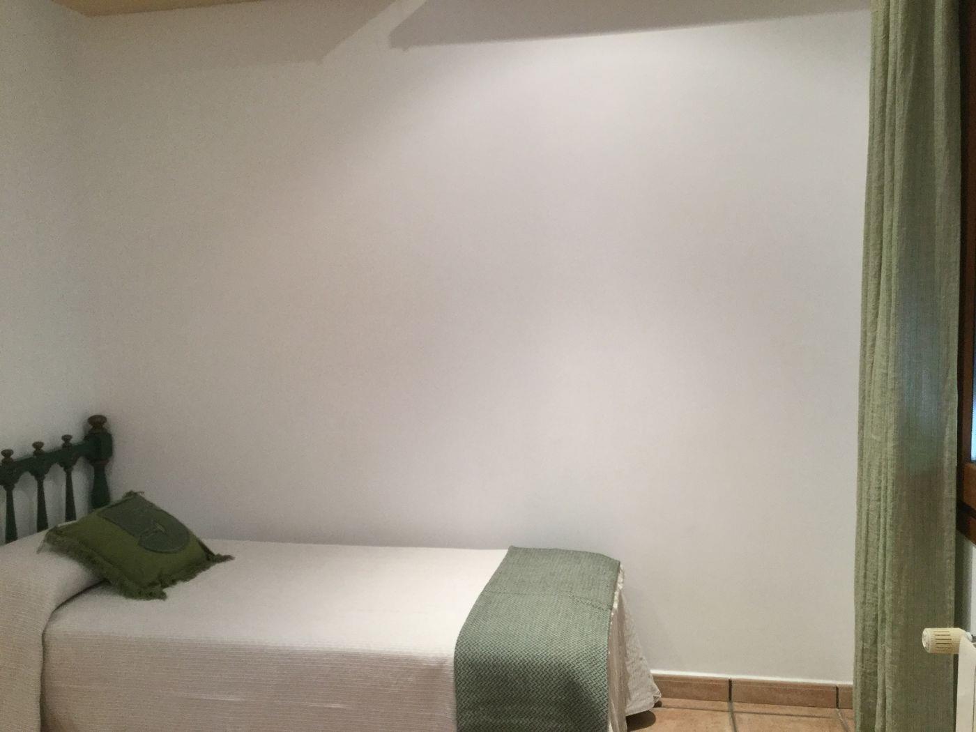 Chambre simple avec tête de lit en bois foncé, literie blanche aux accents verts, sol en terre cuite et rideau vert coordonné.