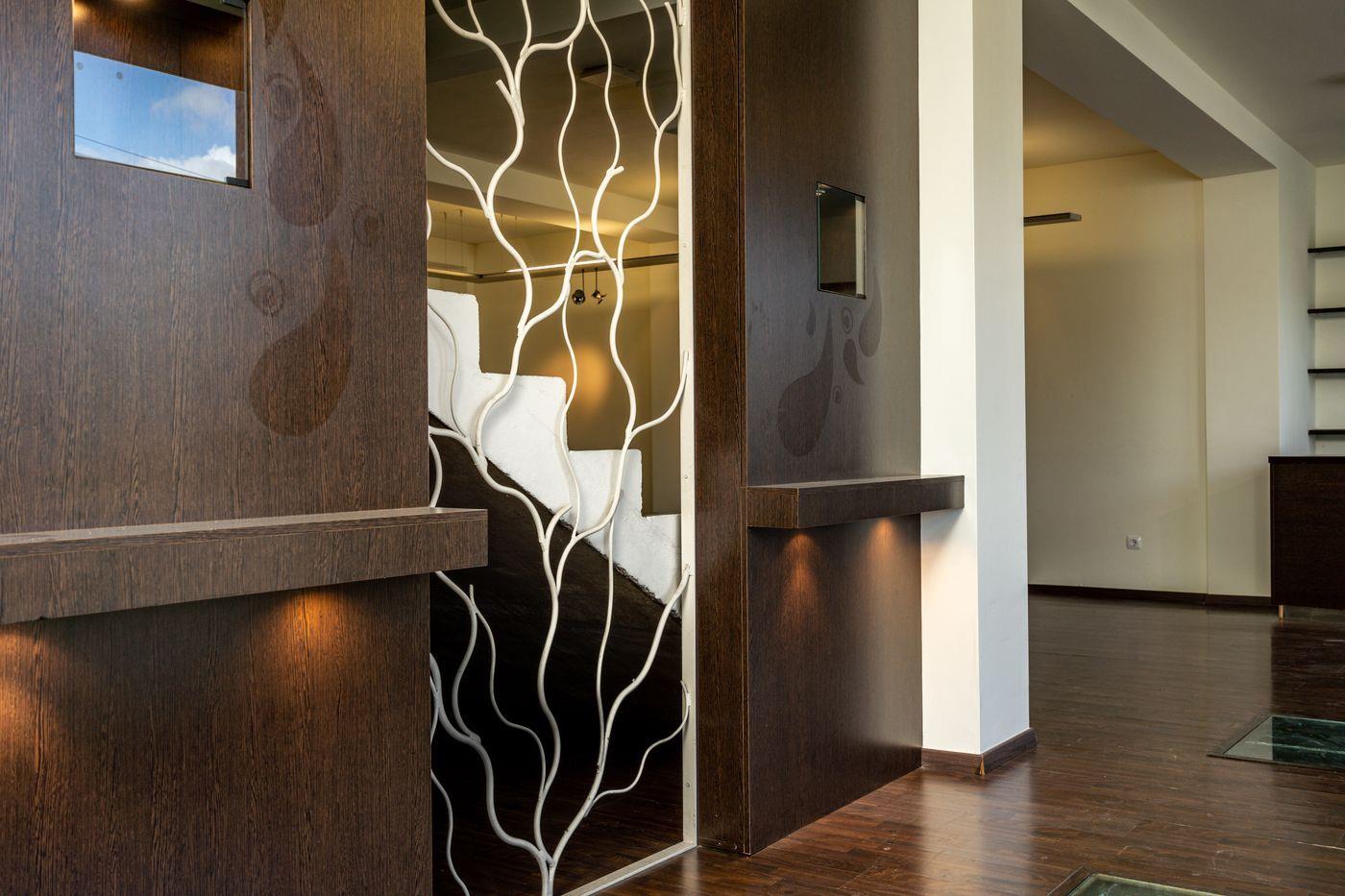 Couloir d'entrée avec boiseries sombres, éclairage intégré et une cloison métallique blanche aux motifs de branches devant l'escalier.