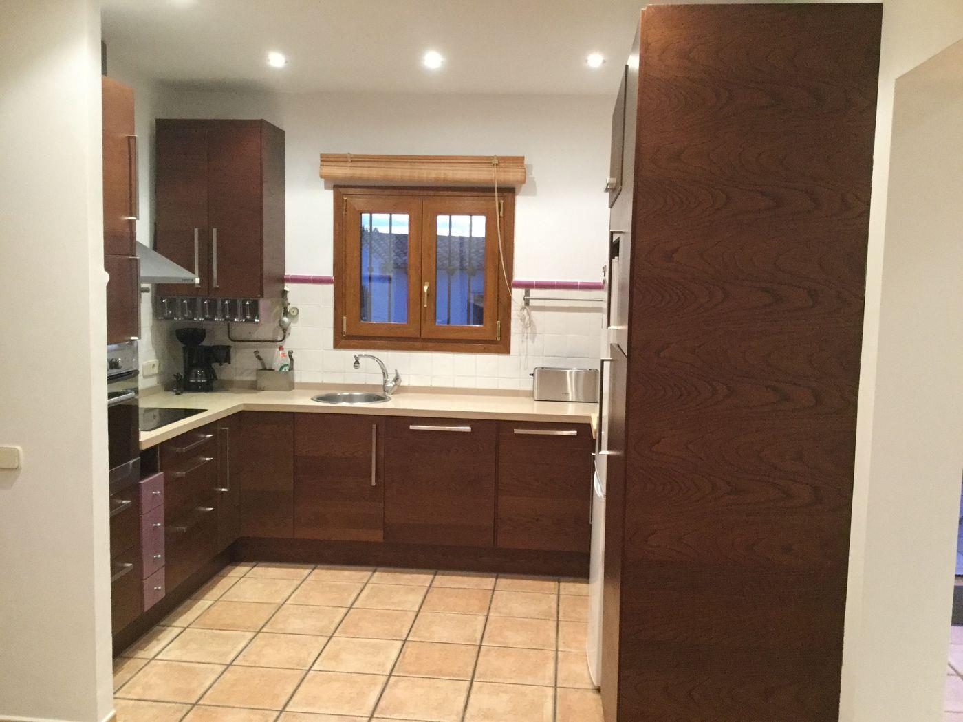 Cuisine équipée avec placards en bois foncé, plan de travail beige et sol en grès cérame. Elle dispose d'une fenêtre à cadre en bois et de spots encastrés.