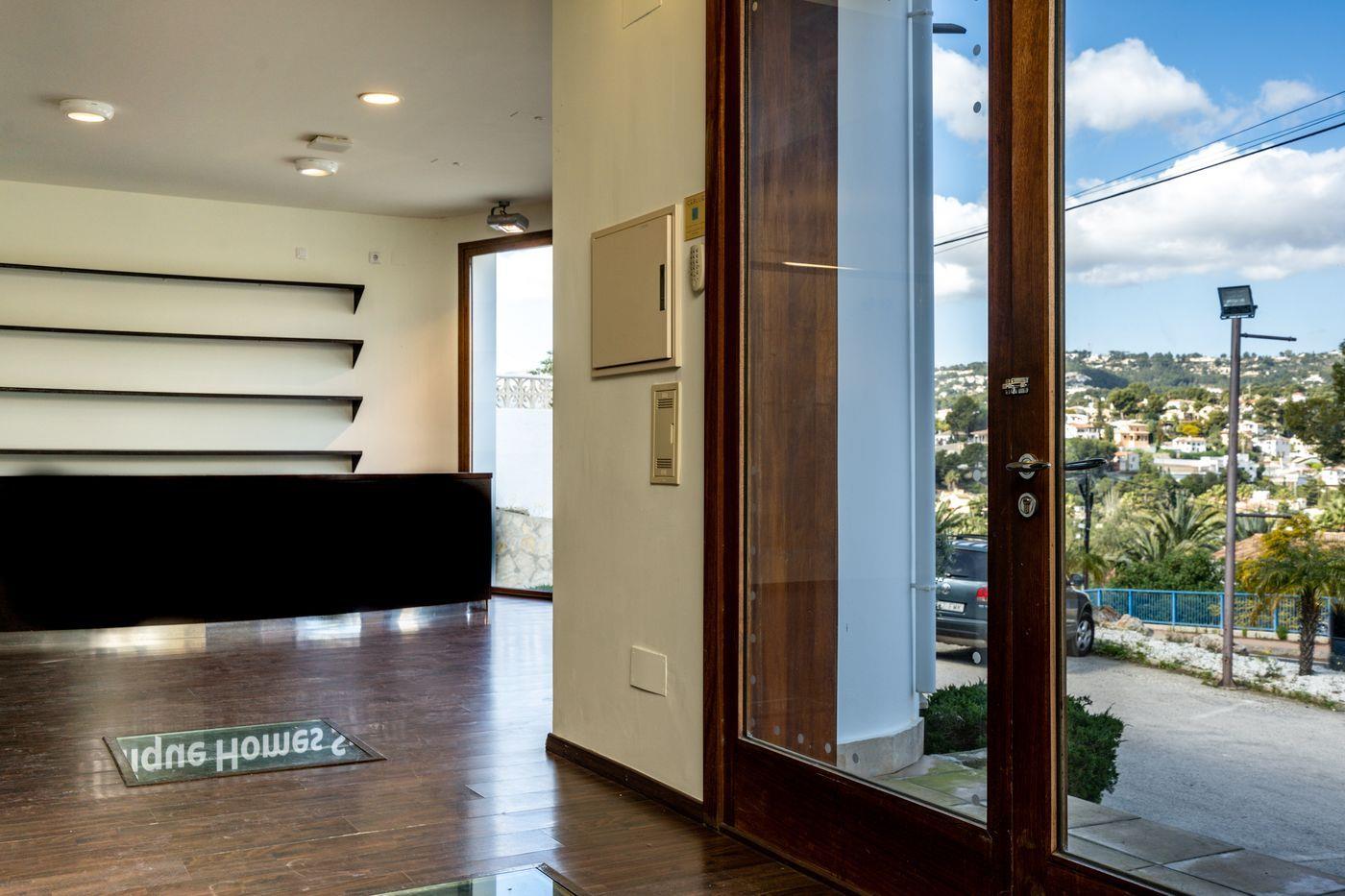 Entrée avec parquet en bois foncé, comptoir de réception noir et porte vitrée. La vue montre un quartier résidentiel vallonné avec des maisons blanches sous un ciel dégagé.