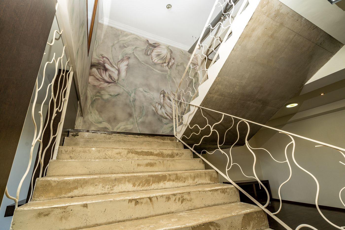 Escalier en béton brut avec garde-corps en fer blanc aux motifs ondulés, mettant en valeur un mur tapissé de tulipes et un éclairage encastré.