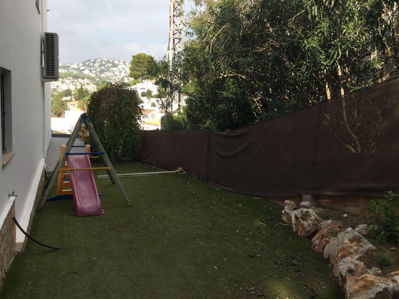 Jardin latéral avec gazon artificiel et aire de jeux pour enfants. L'espace comprend un toboggan et une balançoire, bordés par une clôture d'occultation avec vue sur les collines.