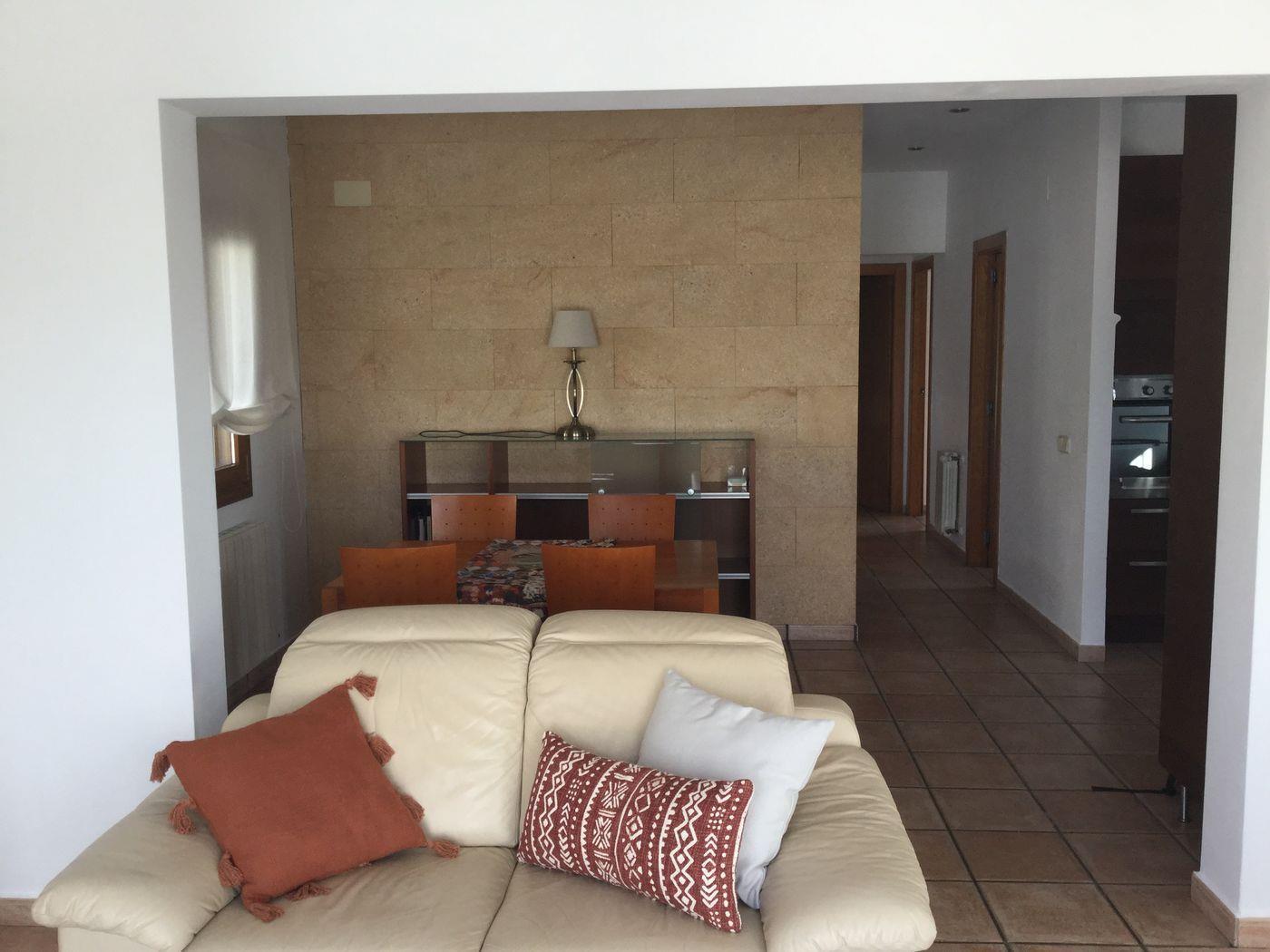 Salon avec canapé en cuir crème et mur d'accent en pierre naturelle. L'espace ouvert mène à une salle à manger et un couloir carrelé vers la cuisine.