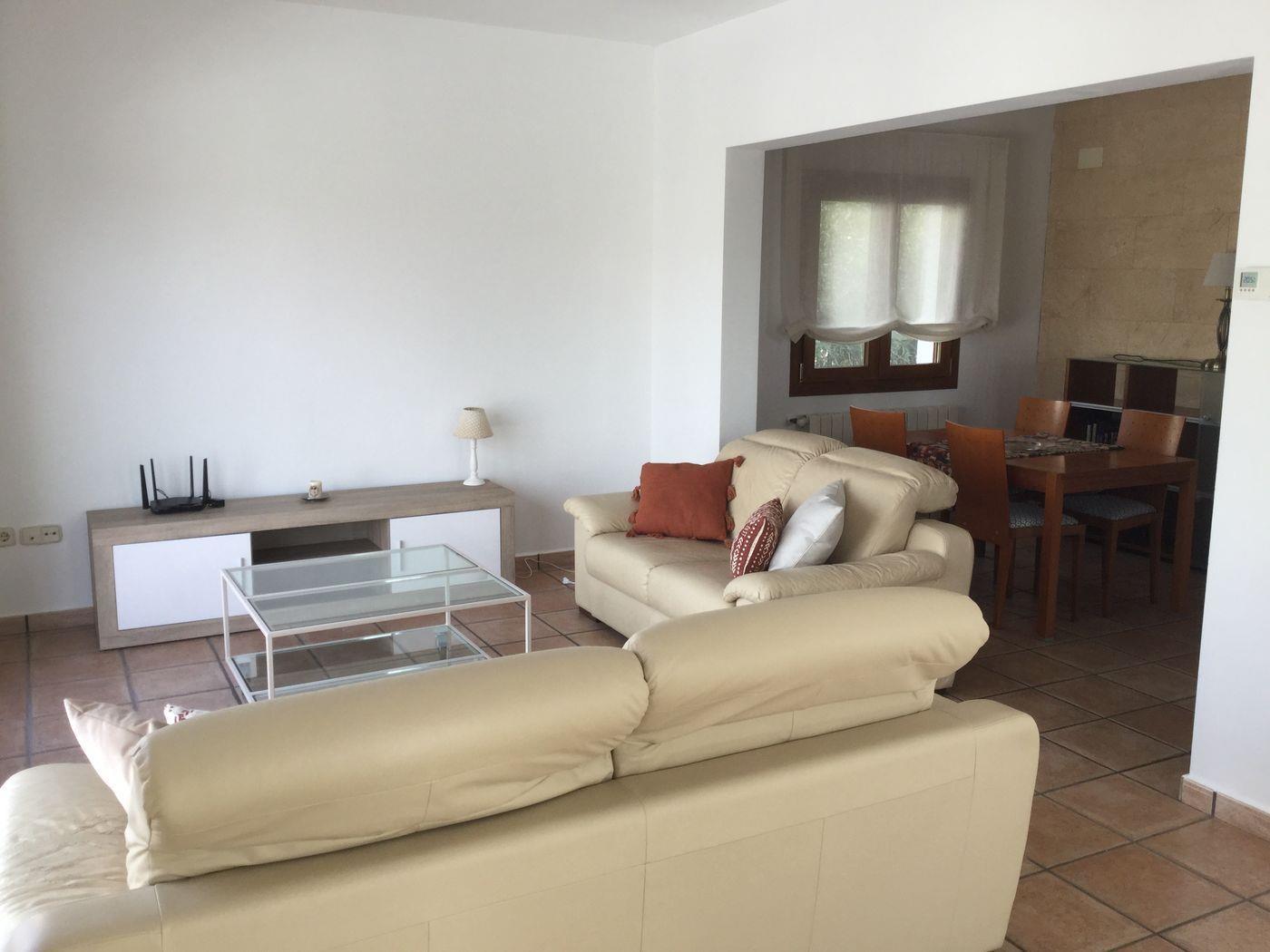 Salon avec canapés en cuir crème, table basse en verre et sol en terre cuite. Espace salle à manger attenant avec un mur de parement en pierre naturelle.