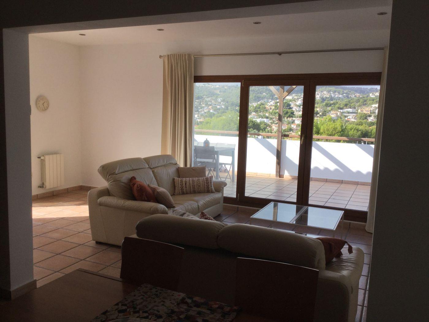 Salon équipé de canapés en cuir crème, sol en terre cuite et grandes baies vitrées ouvrant sur une terrasse avec vue panoramique sur les collines.